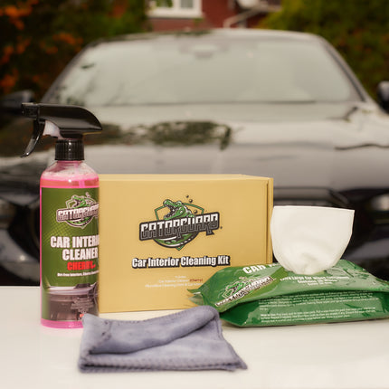 A car interior cleaning kit displayed on a table with a car in the background. The kit includes a bottle of cleaner, a microfiber cleaning cloth, and a box of pre-moistened wipes.